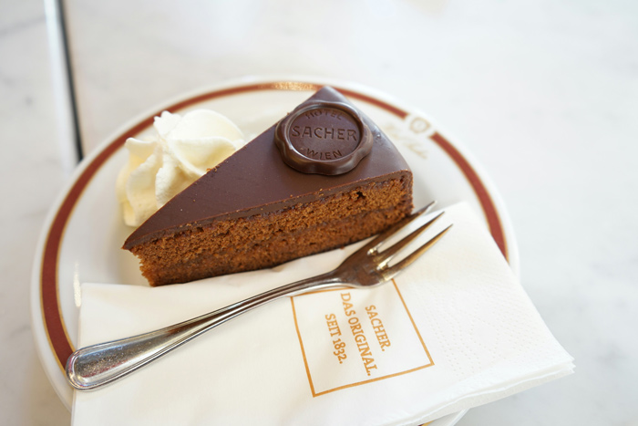 A História da Sachertorte ilustrada por uma fatia do tradicional bolo austríaco, servida em prato clássico com chantilly e selo original do Hotel Sacher.