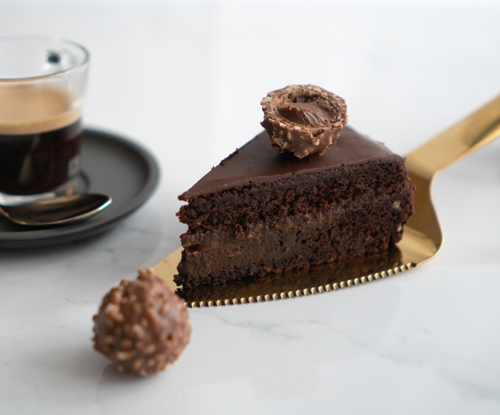 Fatia de Sachertorte com camadas de chocolate brilhante servida em uma pá dourada, acompanhada de um café expresso e um bombom ao fundo.