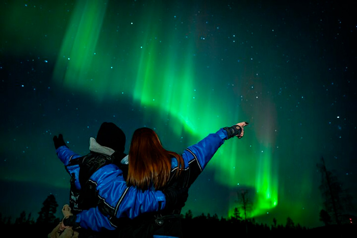 Casal vestindo roupas de frio e apontando para a Aurora boreal na Finlândia, com o céu repleto de estrelas e luzes dançantes ao fundo.