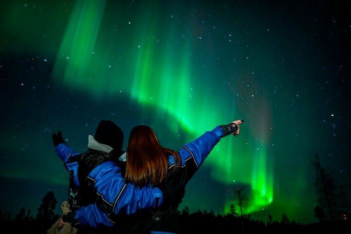 Casal vestindo roupas de frio e apontando para a Aurora boreal na Finlândia, com o céu repleto de estrelas e luzes dançantes ao fundo.