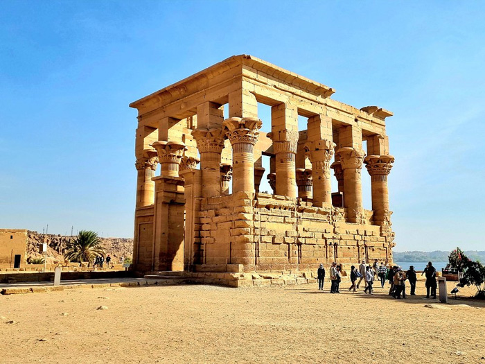 Templo de Philae iluminado pelo sol em Aswan, uma das paradas culturais de um cruzeiro no Nilo, com turistas explorando as ruínas antigas às margens do rio.