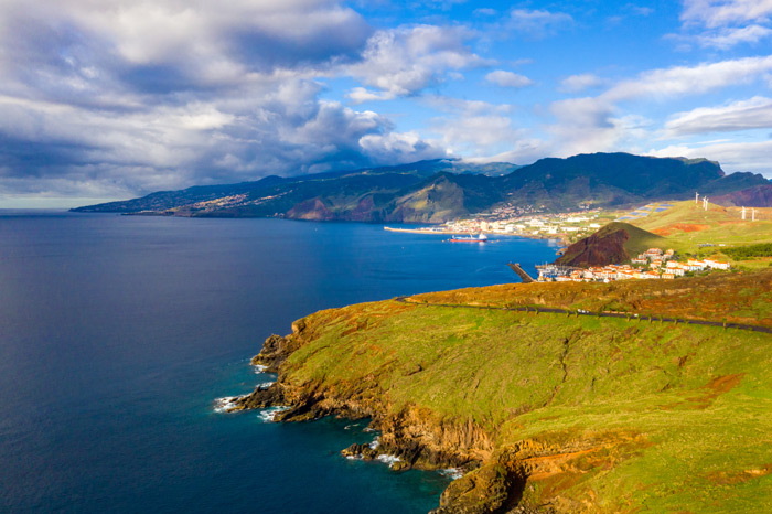 Vista aérea da costa leste da ilha da Madeira, com mar azul profundo, falésias verdes, cidade costeira e montanhas ao fundo sob céu parcialmente nublado. 