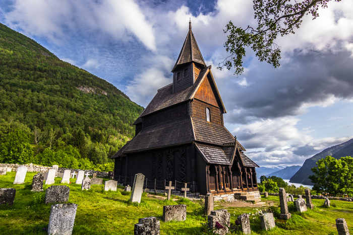 Urnes Stave Church no Sognefjord, igreja medieval de madeira cercada por natureza, com lápides históricas e o fiorde visível ao fundo. 