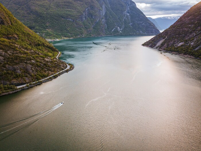 Paisagem ampla do Sognefjord, com águas tranquilas, montanhas ao redor e uma pequena cidade norueguesa refletida no fiorde. 