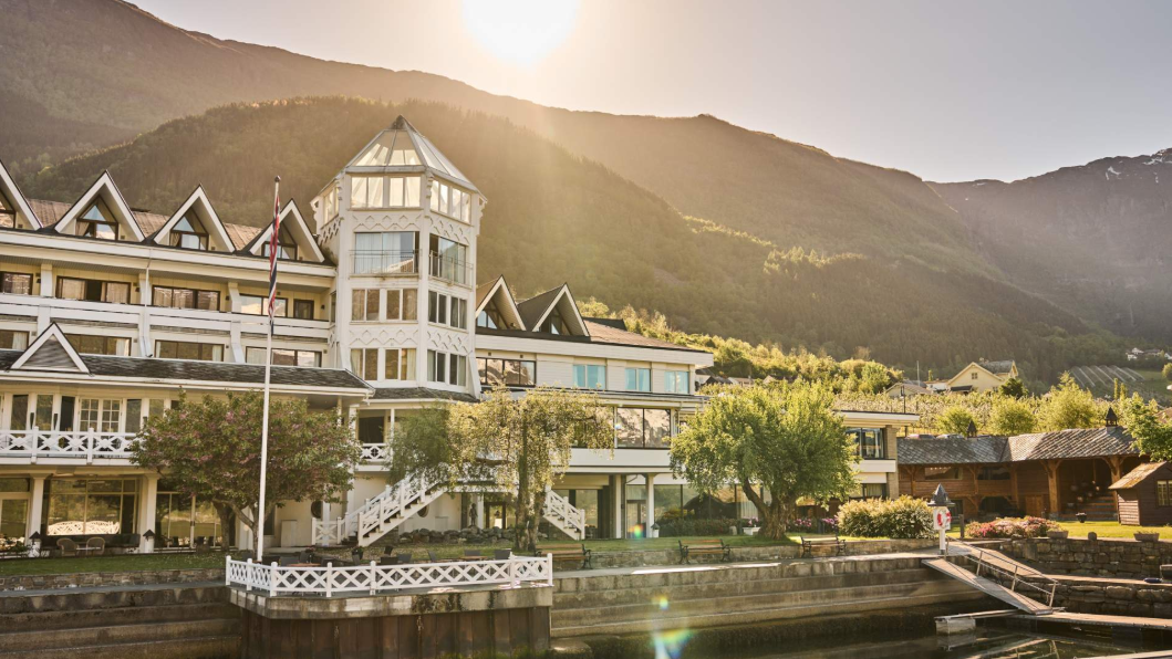 Hotel ullensvang à beira do Sognefjord, com arquitetura clássica norueguesa, varandas de madeira branca, jardim arborizado e montanhas iluminadas pelo sol ao fundo. 