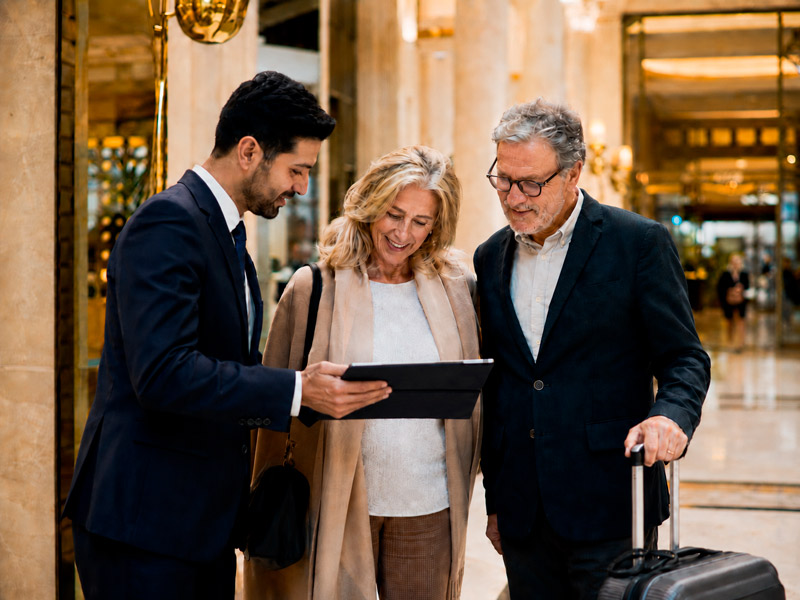 Assistente de viagem auxiliando casal de idosos em viagem internacional, dentro de hotel.