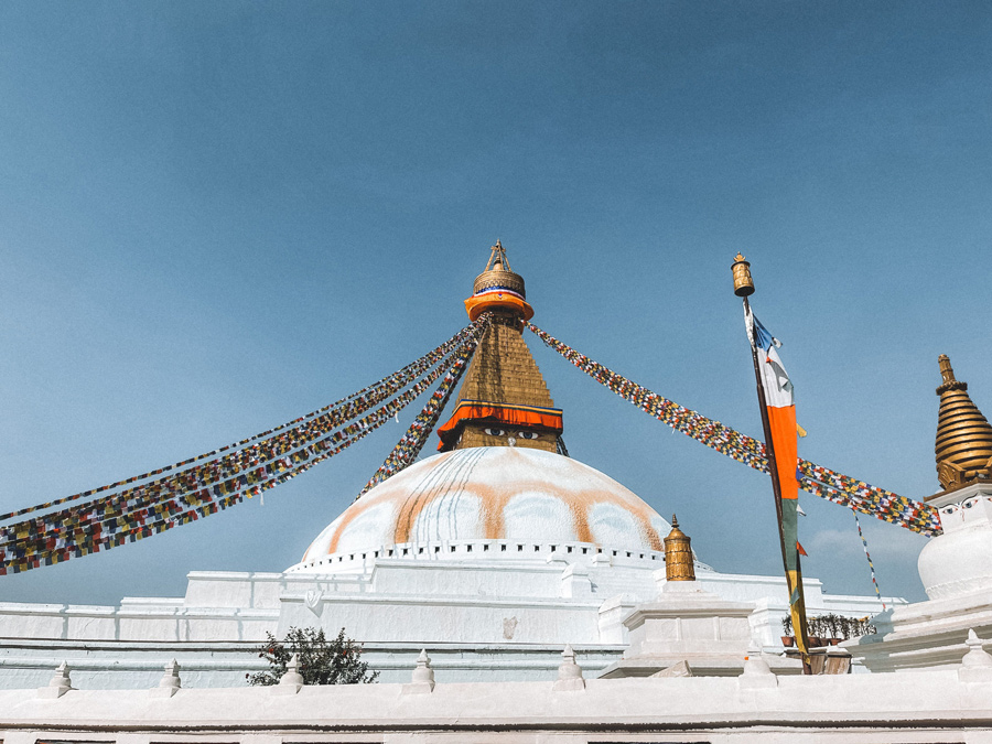 Estupa de Boudhanath, em Katmandu, com a cúpula branca, a torre dourada e bandeiras de oração coloridas contra o céu azul.