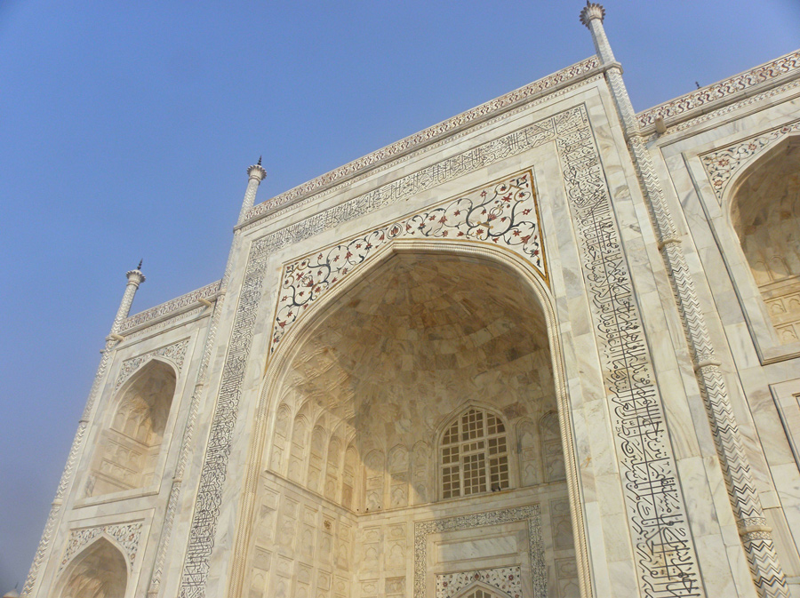 Arco de entrada do Taj Mahal visto de baixo, com inscrições em caligrafia e flores entalhadas no mármore branco.